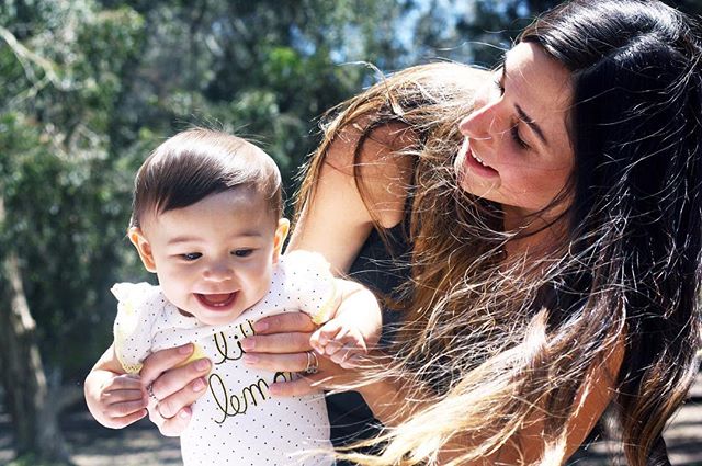 A loving mother holds her smiling baby outdoors in a sun-drenched park setting.