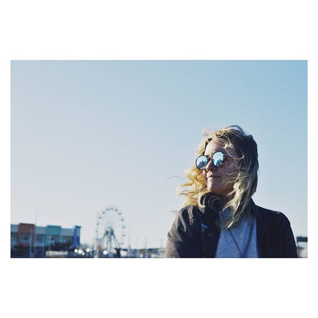 A woman wearing sunglasses and headphones enjoys a sunny day with a ferris wheel in the background.