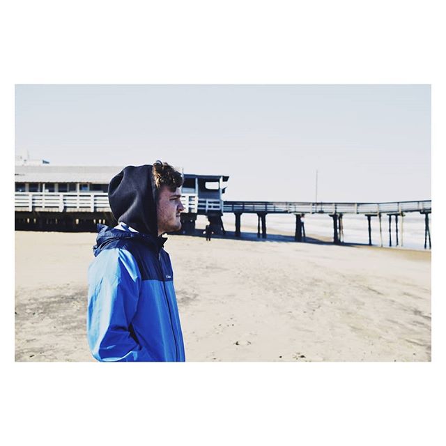 A man in a blue jacket stands on a beach near a pier on a clear day.