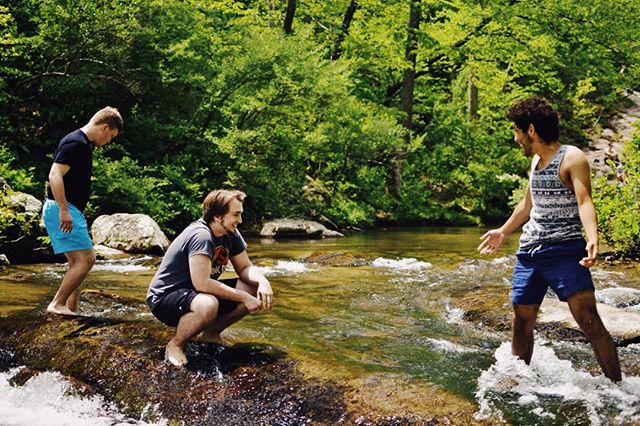 Three friends enjoy wading in a shallow river surrounded by lush greenery on a sunny day.