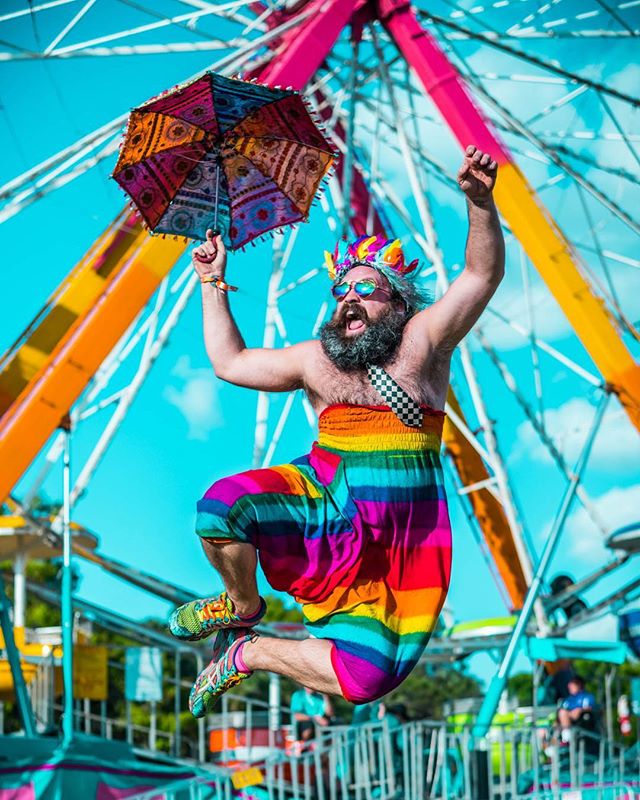 A cheerful man in rainbow attire jumps with joy at a vibrant carnival, holding a colorful umbrella.
