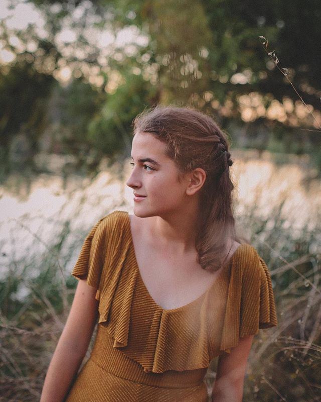 A young woman poses outdoors with a serene expression, bathed in soft, natural light, wearing a golden dress.