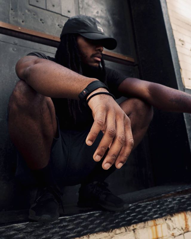A muscular man with dreadlocks sits in an urban setting, showcasing a sporty and athletic style.