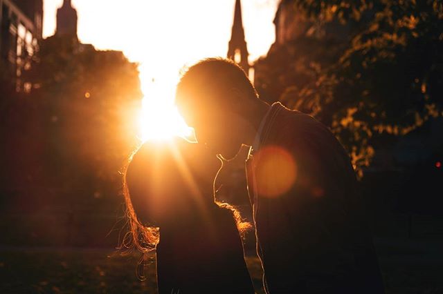 A silhouetted couple shares a tender moment kissing as the sun sets behind them in a city park.