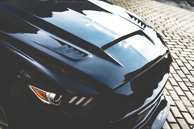 A close-up shot highlights the sleek hood and headlights of a modern black car on a brick street.