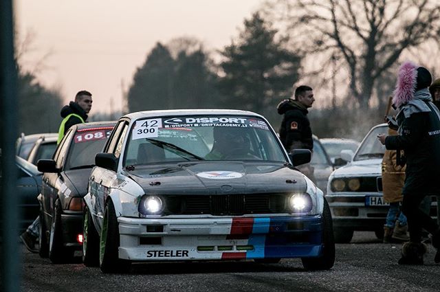A modified BMW E30 race car is parked among people at a motorsport event on a cloudy day.