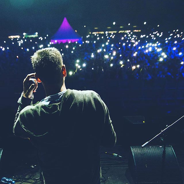 A performer faces a crowd holding lights at an outdoor music festival at night.