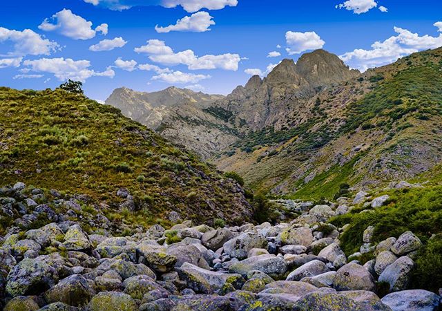 A scenic mountain valley with large rocks, lush greenery, and a bright blue sky filled with fluffy white clouds.