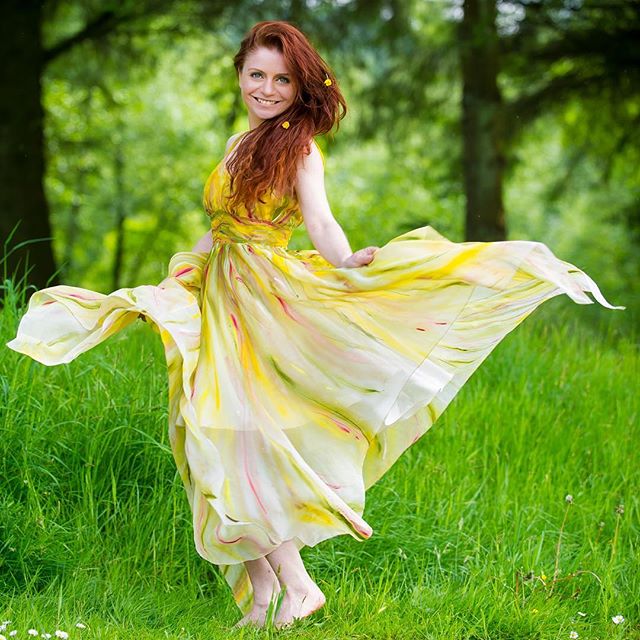 A vibrant woman in a yellow dress dances joyfully in a lush, green meadow surrounded by trees, embodying the essence of spring.