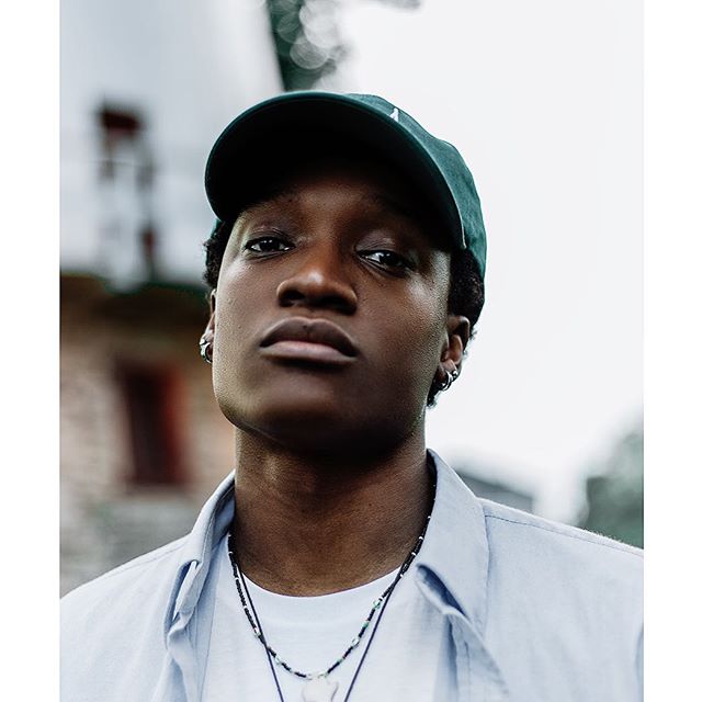 A young man with a green cap poses in an urban setting, wearing a light blue shirt and necklace.