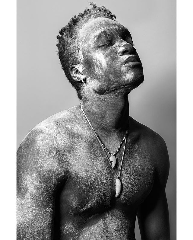 A black and white studio shot of a man with a necklace, eyes closed, head tilted back.