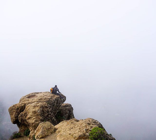 A lone hiker sits atop a rocky peak, enjoying the view amidst the misty mountain landscape.