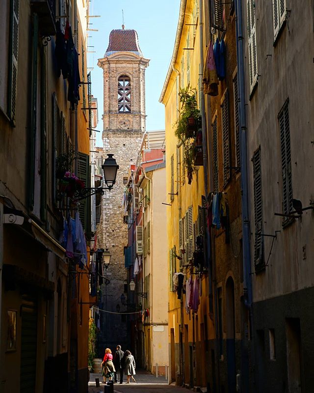 People walk down a narrow street with a bell tower in the distance, capturing the essence of an old city.