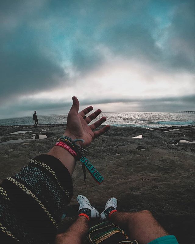 Person's perspective at the beach, hand outstretched under a cloudy sky, promoting relaxation and outdoor lifestyle.