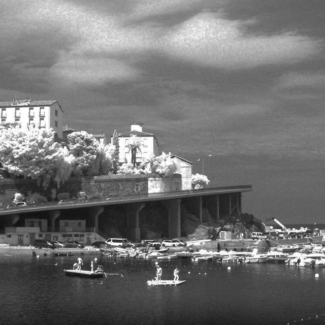 A black and white image of a waterfront town with buildings, boats, and people enjoying the water.