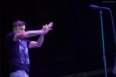 A tattooed singer performs on stage with a microphone in front of a dark backdrop.