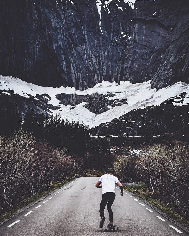 A skateboarder rides down a mountain road, surrounded by stunning snowy peaks and dense trees.