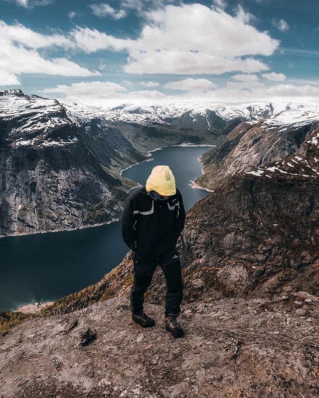 A lone hiker stands atop a rocky mountain, overlooking a serene fjord landscape in Norway.