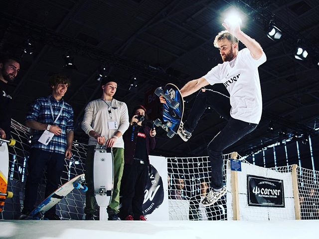 A skateboarder performs a trick at an indoor skatepark, surrounded by onlookers at an event. 