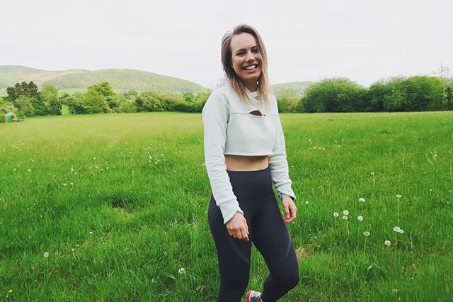 A smiling woman in athletic wear stands in a sunny meadow, promoting fitness and an active outdoor lifestyle.