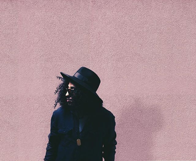 A stylish man with curly hair and a hat stands in front of a pink wall, looking away.