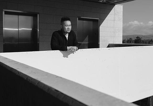 A man thoughtfully gazes out from a balcony in a black and white urban setting.