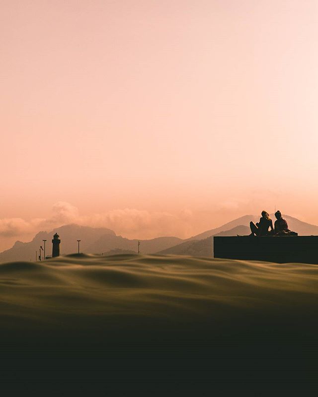 A silhouette of two people relaxing near a lighthouse on a sandy beach against a dreamy, warm-toned sunset.