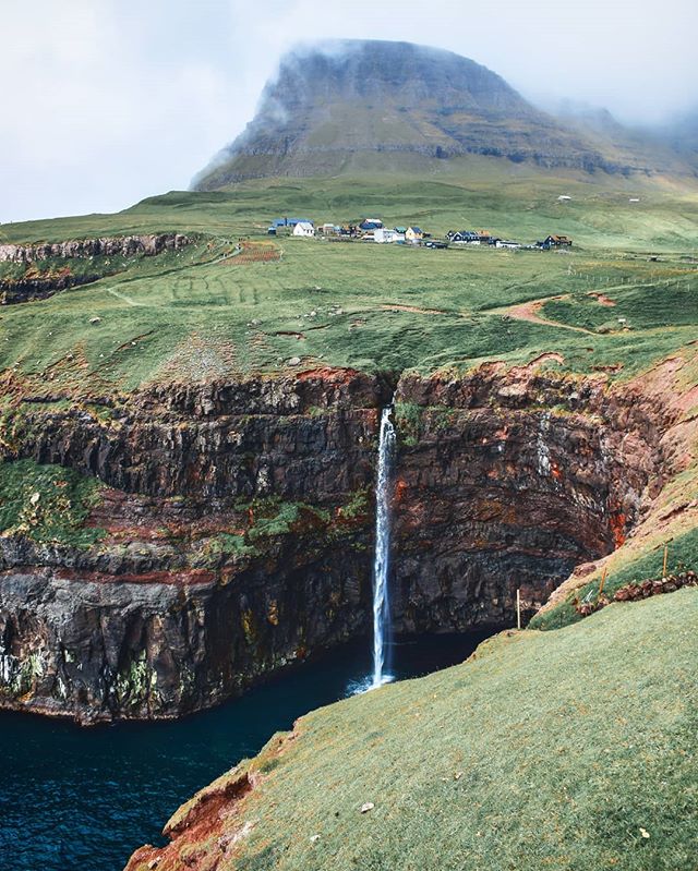 A scenic waterfall flows into the ocean near a village beneath a green mountain.