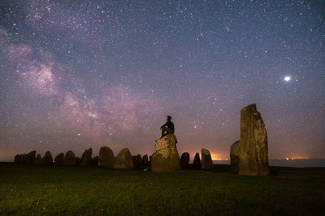 A lone man sits among ancient stones under a starlit sky, gazing at the Milky Way.