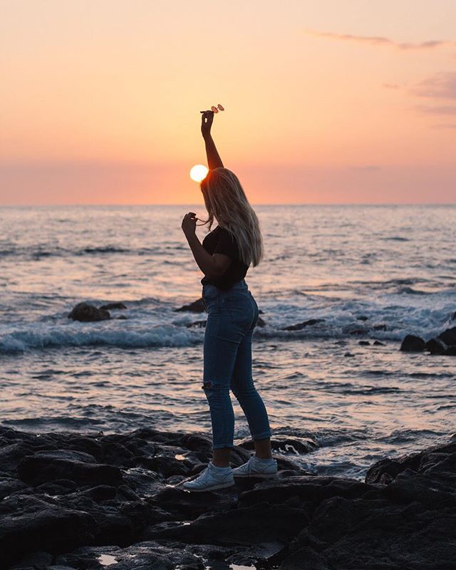 A woman silhouetted against the sunset stands on a rocky beach, holding sunglasses aloft.