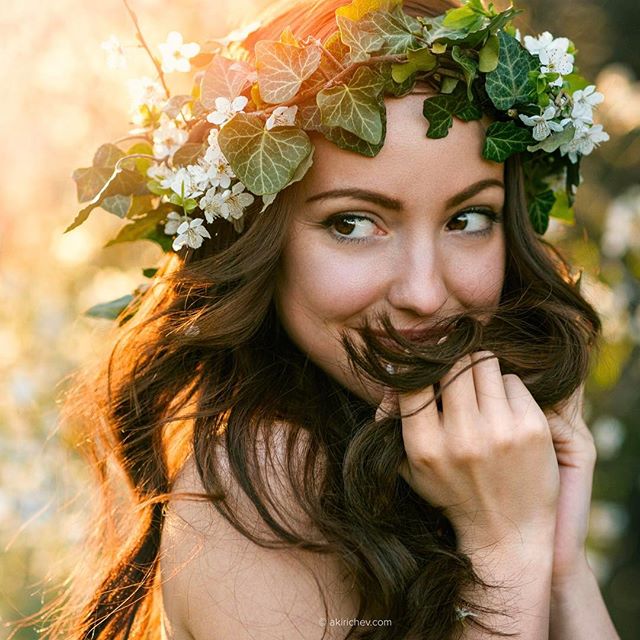 A cheerful woman with a flower crown smiles dreamily in a soft, natural outdoor setting.