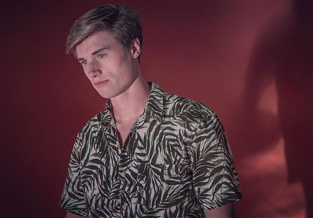 A young man in a patterned shirt poses against a red backdrop in a studio setting.