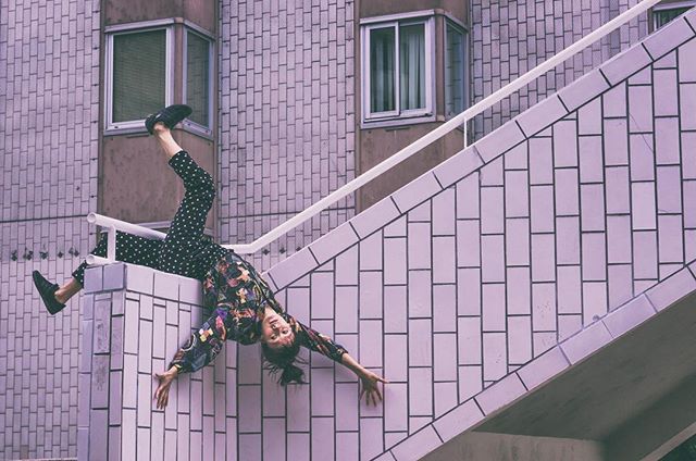 A woman in patterned clothing playfully poses upside down on outdoor stairs against a building.