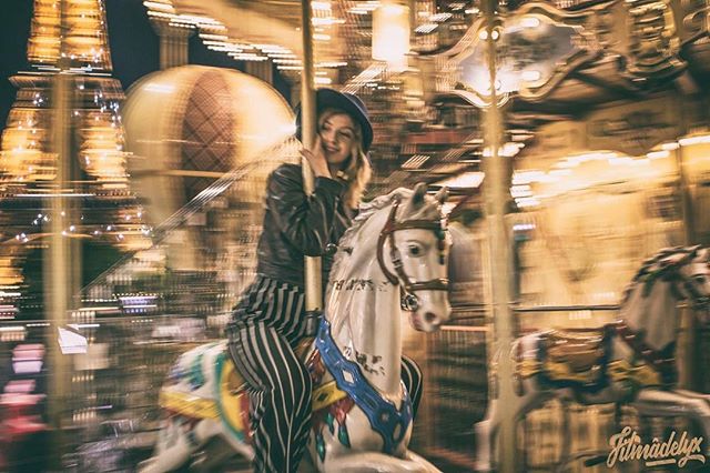 A woman enjoys a carousel ride at night, wearing striped pants and a hat, bathed in warm, muted light.