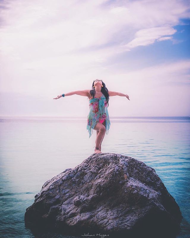 A woman stands on a rock by the ocean with arms outstretched, enjoying a peaceful moment in a serene setting.