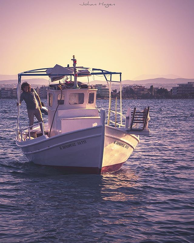 A man stands on a boat, enjoying the view of the cityscape and calm sea at sunset.