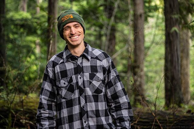 A smiling man in a plaid shirt and hat stands in a forest, enjoying the outdoors.