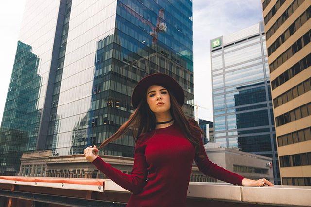 A confident woman in a hat poses in an urban setting with modern buildings for a fashion shoot.