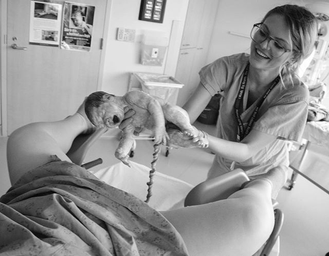 A nurse smiles holding a newborn baby with the umbilical cord in a hospital room after delivery.