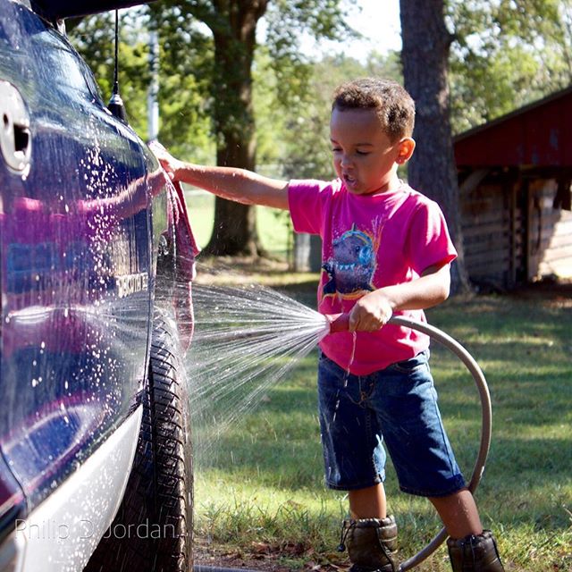A young boy smiles as he washes a car with a hose outside on a sunny day.