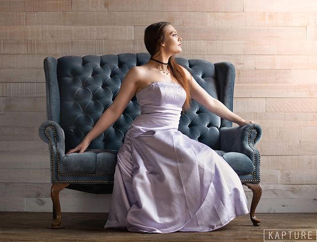 A woman in a lilac dress sits in an elegant armchair in a studio setting.