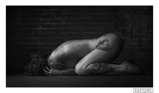 A nude woman with tattoos poses artistically against a brick wall in a black and white studio setting.