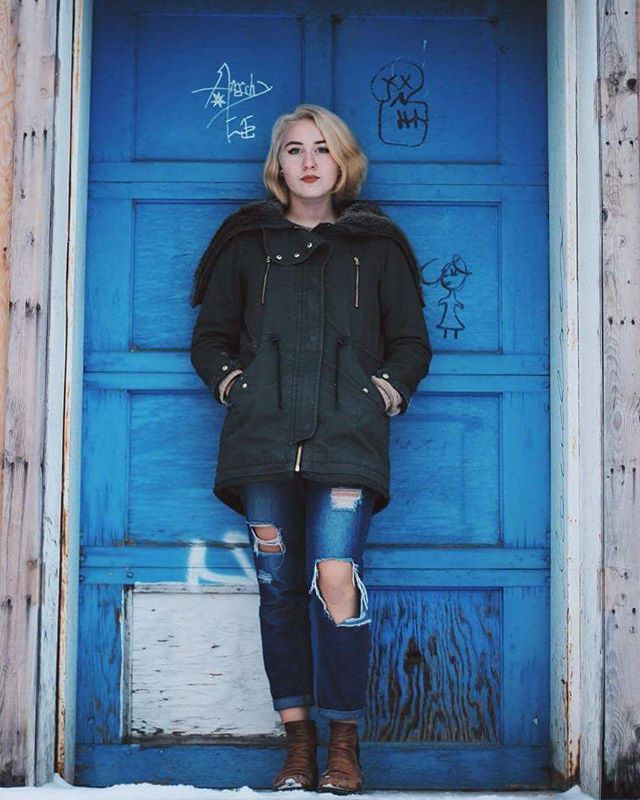 A woman poses in front of a blue door in ripped jeans and a jacket.