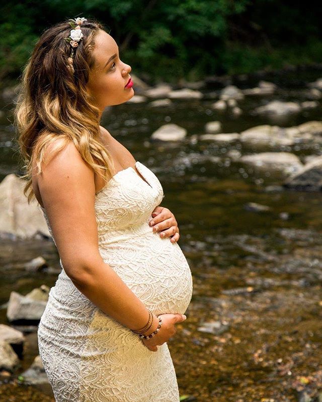 A pregnant woman in a white dress poses by a river, exuding serene beauty and anticipation.