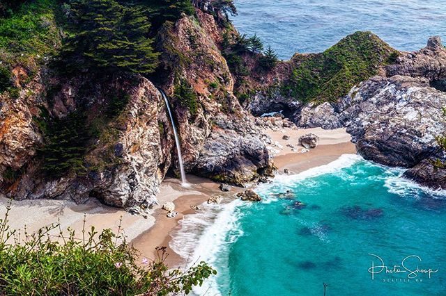 A picturesque waterfall cascades onto a sandy beach, meeting the turquoise ocean along a rugged coastline.