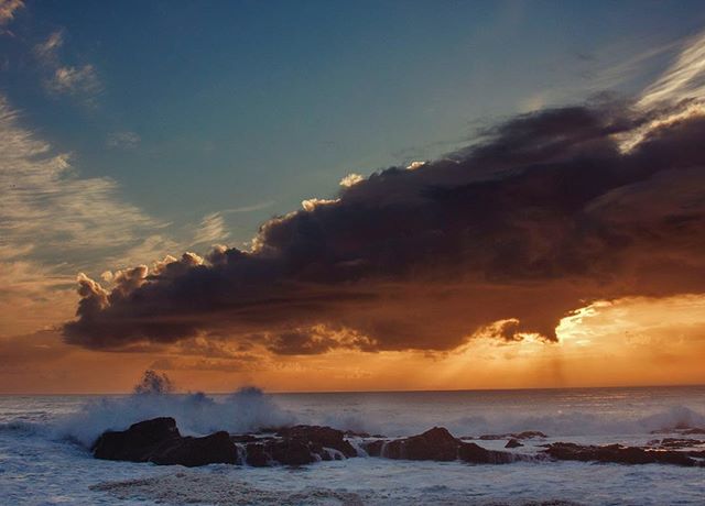 Waves crash against the rocky shore at sunset, with dramatic clouds in the sky.