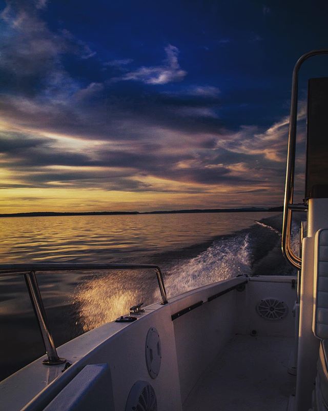 A boat sails on the water during sunset, with dramatic clouds above. Water spray marks the wake.