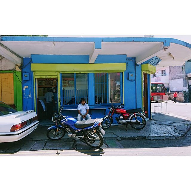 A street scene with a man sitting outside a colorful building with motorcycles parked nearby and a car on the side.
