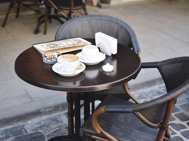 A cozy outdoor cafe scene with a coffee cup, menu, and ashtray on a round wooden table with chairs.