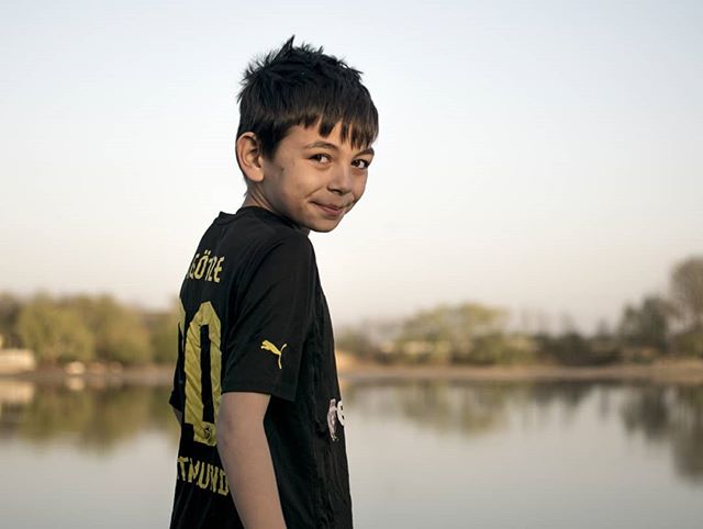 A smiling boy in a black soccer jersey stands near a lake, looking back over his shoulder on a bright day.